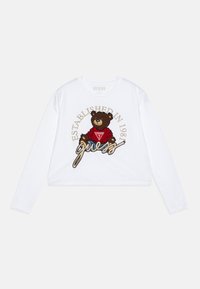 Weißes langärmliges Cropped-Top mit einem Grafikdruck eines braunen Bären in einem roten Shirt, versehen mit der Aufschrift "GUESS" und "ESTABLISHED IN 1981" in goldener Stickerei.