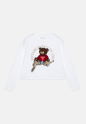 Weißes langärmliges Cropped-Top mit einem Grafikdruck eines braunen Bären in einem roten Shirt, versehen mit der Aufschrift "GUESS" und "ESTABLISHED IN 1981" in goldener Stickerei.