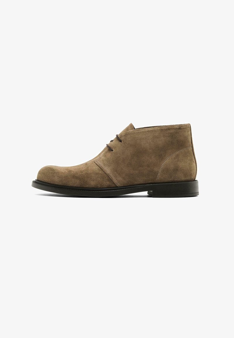 Stivali chukka in suede marrone con punta rotonda, due occhielli per lacci e suola piatta in gomma. La scarpa presenta una texture liscia e un design minimalista.