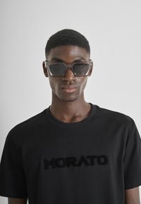 Gafas de sol con un marco gris transparente y lentes oscuros, combinadas con una camiseta negra que presenta un logo en relieve de "MORATO".