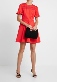 Robe rouge avec des manches courtes à volants, corsage texturé, jupe évasée, accompagnée d'un sac à main en suédine noire avec une poignée dorée.