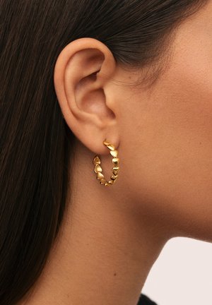 Primo piano di un orecchio con un tono di pelle medio che indossa un orecchino a cerchio d'oro composto da piccoli link a forma di cuore. Capelli castano scuri visibili.