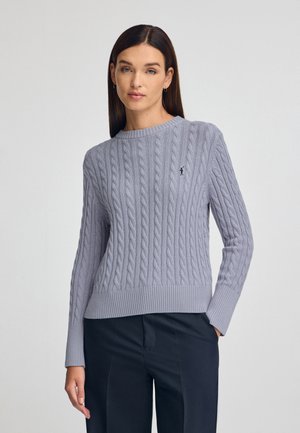 Hellgrauer, kabelgestrickter Pullover mit Rundhalsausschnitt, gerippten Bündchen und Saum, versehen mit einem kleinen schwarzen Logo auf der Brust. Getragen mit dunklen Hosen.