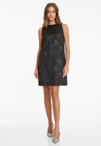Frau, die ein ärmelloses schwarzes knielanges Kleid mit floralen Mustern und silbernen High Heels trägt, vor einem weißen Hintergrund steht.
