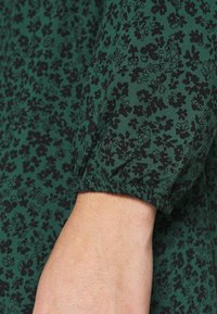 Tissu de blouse florale verte avec un motif floral noir, présentant un détail de poignet élastique au niveau des manches. Texture lisse, matériau léger.