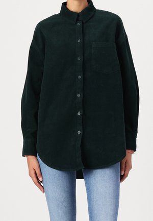 Tummanvihreä pitkähihainen puuvillapaita, jossa nappikiinnitys ja edessä tasku, päällä vaaleansiniset farkut seisovalla henkilöllä.