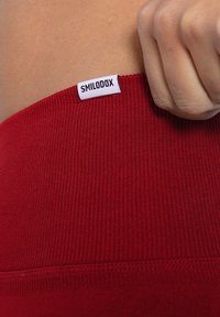 Rood geribbeld stof met een zichtbaar wit label met de tekst "SMILODOX." Close-up weergave richt zich op de tailleband en textuurdetails.