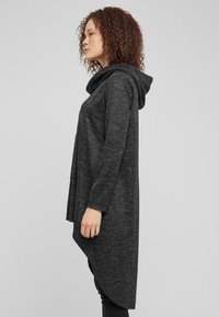 Svart hettegenser med teksturert strikk, med asymmetrisk hem og lange ermer. Materialet har en myk finish.