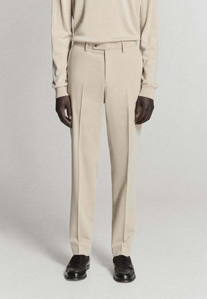 Mann trägt beige plissierte Hose, passendes beige langärmeliges Oberteil und schwarze Ledermokassins, steht vor einem neutralen Hintergrund.