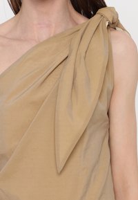 Gros plan sur une personne portant un haut beige asymétrique avec un grand nœud noué sur l'épaule gauche.