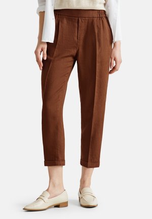 Femme portant un pantalon marron fuselé longueur cheville avec ourlets retroussés et mocassins crème sans lacets, montrée de la taille aux pieds.