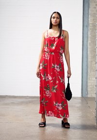 Vestido maxi vermelho com padrão floral, cintura elástica, alças ajustáveis e bolsa de tecido preto. Modelo a usar sandálias pretas.