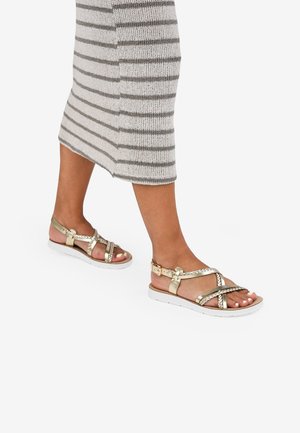Sandalias doradas con tiras, diseño trenzado y hebilla ajustable, montadas sobre una suela blanca. Llevadas con un vestido de punto a rayas en gris.