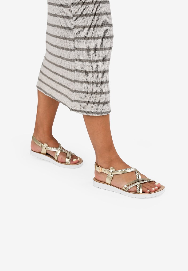 Sandalias doradas con tiras, diseño trenzado y hebilla ajustable, montadas sobre una suela blanca. Llevadas con un vestido de punto a rayas en gris.