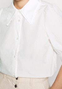 Hvid kortærmet skjorte med en stor, flæset krave og knaplukning. Fremstillet af letvægtsstof og har en glat tekstur.