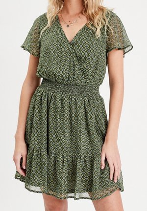 Vrouw die een groen jurk met korte mouwen draagt, bedrukt met een patroon, met een V-hals, elastische tailleband en een gerimpelde zoom, staand tegen een effen achtergrond.
