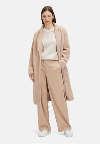 Lang beige jas van textuurstof, gedragen over een licht sweater en beige corduroy broek, gecombineerd met witte sneakers.
