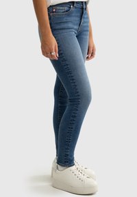 Keskmise sinise denimi kitsad teksased ühtlaset tekstuuriga, klassikalise viie taskuga disain ja kontrastne õmblus. Selga pandud valged spordijalatsid.