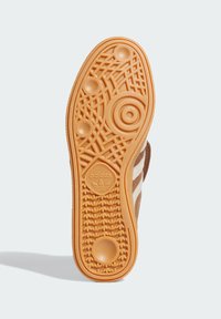 Podrážka hnedých adidas tenisiek s gumovou podošvou, vzorovaným dezénom, logom adidas trefoil a bielymi pruhmi viditeľnými po stranách.