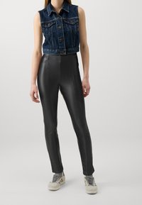 Denim ärmlös jacka, mörkblå, med frontknappar och bröstfickor; matchad med figursydda svarta konstläderbyxor och ljusa sneakers.