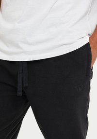 Zwarte fleece joggingbroeken met een trekkoord in de taille en zijzakken. Subtiele, reliëf logo-detail op het been. Gepaard met een wit T-shirt.