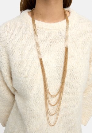 Długi złoty naszyjnik warstwowy noszony na kremowym, teksturowanym swetrze na osobie.