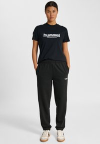 Sort Hummel T-shirt med hvidt logo, parret med sorte sweatpants med logo og lys sneakers med beige og sorte accenter.