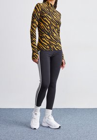 Dlouhý rukáv košile v žlutočerném zvířecím vzoru, doplněný černými legínami s bílými bočními pruhy a bílými sportovn ími botami.