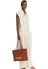 Gilet beige senza maniche con cerniere frontali, abbinato a pantaloni ampi color beige chiaro. Borsa tote marrone con logo "LOVE MOSCHINO".