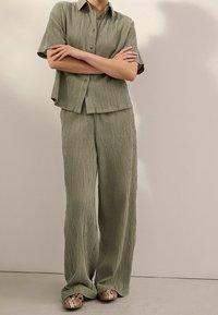 Olive green textured shirt and pants set featuring a relaxed fit, button closure, and visible pleating; paired with patterned slide shoes.