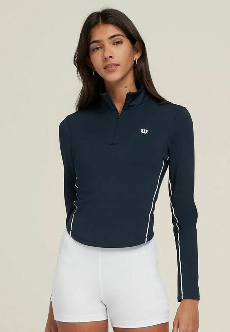 Top a maniche lunghe blu navy con colletto a metà zip, strisce bianche sui lati e un logo discreto sul petto. Il tessuto appare liscio ed elasticizzato.