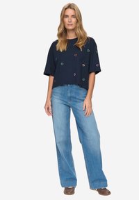 Camiseta corta azul marino con bordado de corazón multicolor, combinada con vaqueros de pierna ancha en azul claro. Tela casual y ajuste relajado.