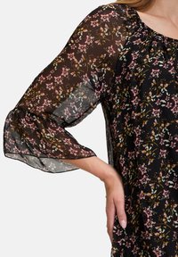 Musta kukallinen bluuza läpinäkyvillä hihoilla, jossa on vaaleanpunainen ja vihreä kukkakuosi, röyhkeä kaula-aukko ja rento istuvuus.