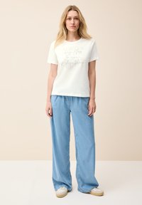 Vit bomull t-shirt med tryckt text i silver, i kombination med ljusblå vida jeans och vita skor; enkel design och avslappnad passform.