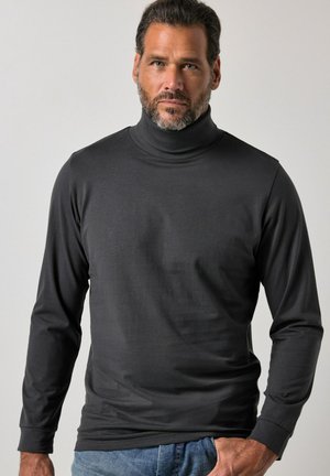 Homme barbu portant un pull roulé noir à manches longues et un jean bleu, debout devant un fond clair uni.