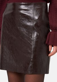 Mini gonna in finta pelle marrone con finitura lucida, caratterizzata da una silhouette dritta, cuciture a vista e una texture liscia.