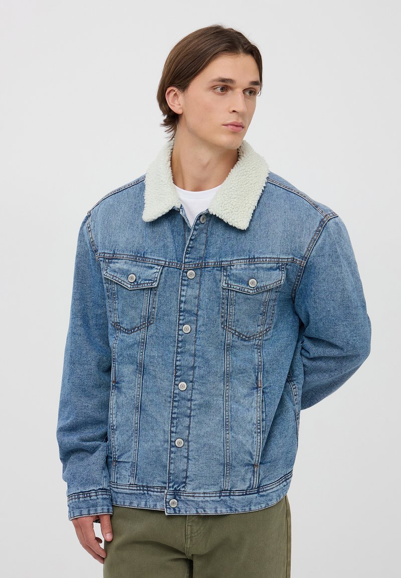 Giubbotto in denim di un azzurro chiaro con colletto in sherpa bianco, dotato di due tasche sul petto e bottoni a pressione argentati. Risulta avere una texture liscia e una vestibilità casual.