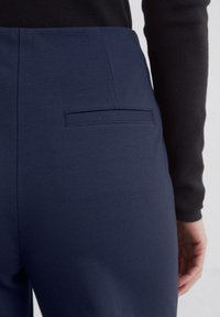 Pantaloni navy blue, croiți, cu o textură netedă, având un singur buzunar la spate și un design cu talie înaltă.
