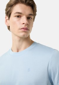 Světlomodré tričko z měkké látky, s kulatým výstřihem a vyraženým logem ve tvaru písmena "B" na hrudi.