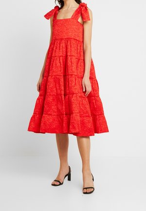 Woman wearing a knee-length, tiered red dress with bow shoulder straps and textured floral fabric, paired with black high-heeled sandals.
