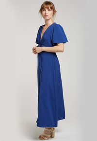 Robe longue bleue en tissu lisse. Présente un décolleté en V profond, des manches courtes et flottantes, ainsi qu'un lien à l'avant. Comprend une fente latérale discrète.