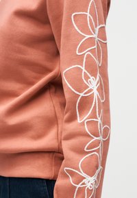 Χέρι με πουλόβερ σε ξεθωριασμένο ροζ χρώμα με λευκές κεντητές λουλούδια, συνδυασμένο με σκούρο μπλε τζιν μπροστά σε ανοιχτό φόντο.