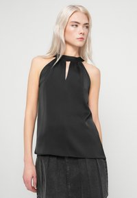 Jovem mulher com cabelo loiro platinado, vestindo um top preto sem mangas e uma saia de ganga cinza escura, em frente a um fundo simples.