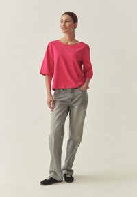 Haut à manches courtes rose vif avec un large décolleté, associé à un jean évasé gris clair et des chaussures noires, debout devant un fond neutre.