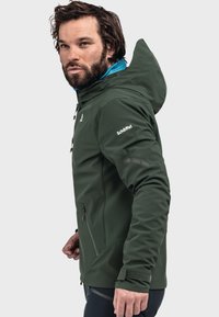 Grüne Softshell-Jacke mit blauer Innenseite, mit Reißverschlusstaschen, verstellbarer Kapuze und Klettverschluss-Manschetten, mit dem Markenlogo auf dem Ärmel.