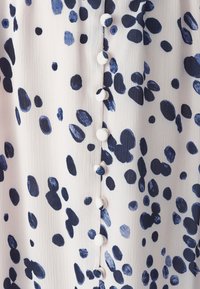 Tissu à motif floral en beige clair avec des taches irrégulières bleu marine. Présente une patte de boutonnage avec des boutons assortis sur le devant.