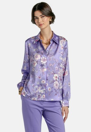 Nő levendulakék virágos szatén blúzban, melyhez passzoló nadrágot, aranylánc nyakláncot és fülbevalókat visel, fehér háttér előtt áll.