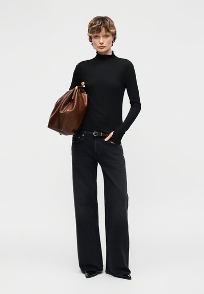 Γυναίκα με μαύρο πλεκτό ζιβάγκο και φαρδιά μαύρα τζιν παντελόνια που κρατά μεγάλη καφέ δερμάτινη τσάντα με χρυσό κούμπωμα.