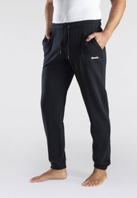Pantalon de jogging noir avec une taille élastique, un cordon de serrage, deux poches latérales et un logo "Bench" blanc sur la poche gauche. Matériau doux.