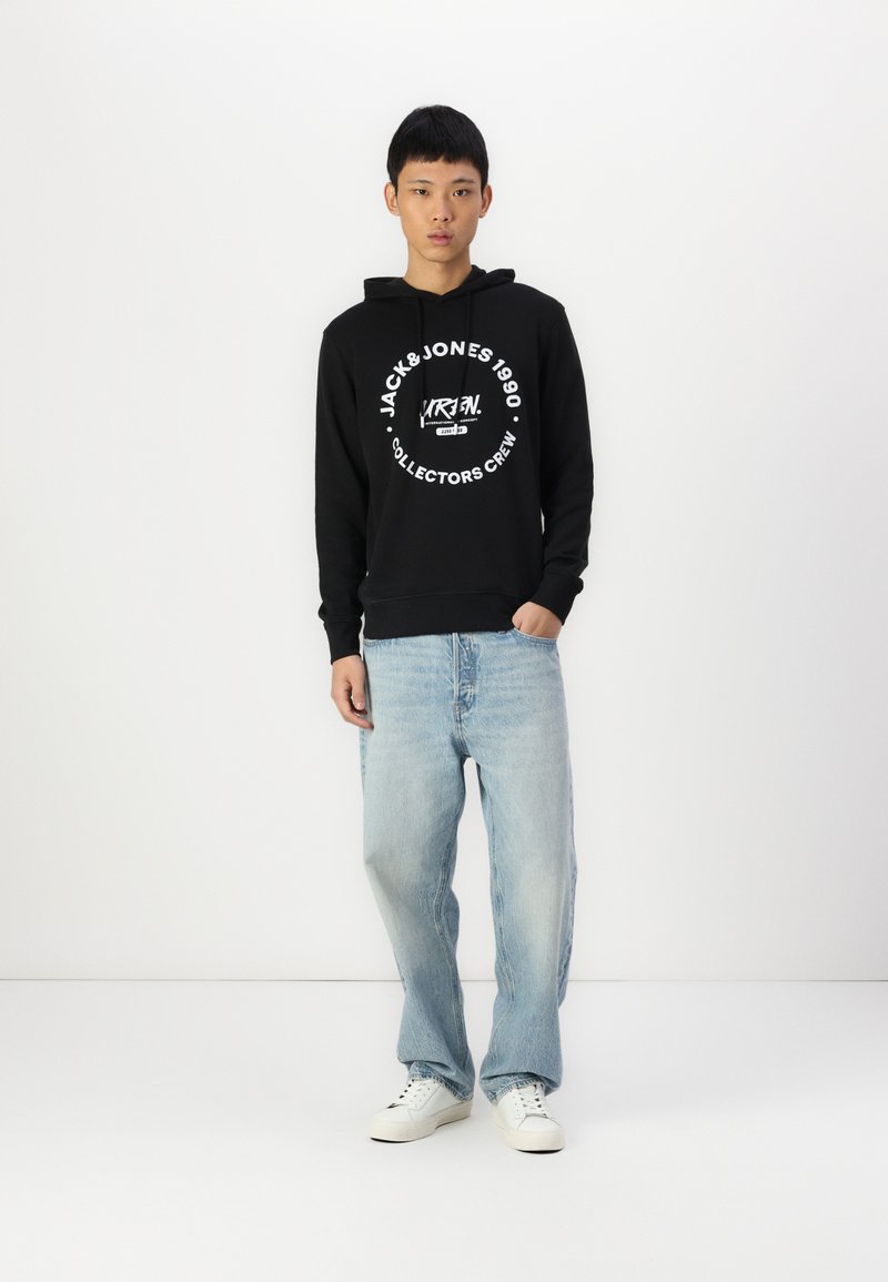 Schwarze Kapuzenjacke mit weißem Text, kombiniert mit hellblauen, lässig sitzenden Jeans und weißen Sneakers, vor einem schlichten Hintergrund stehend.